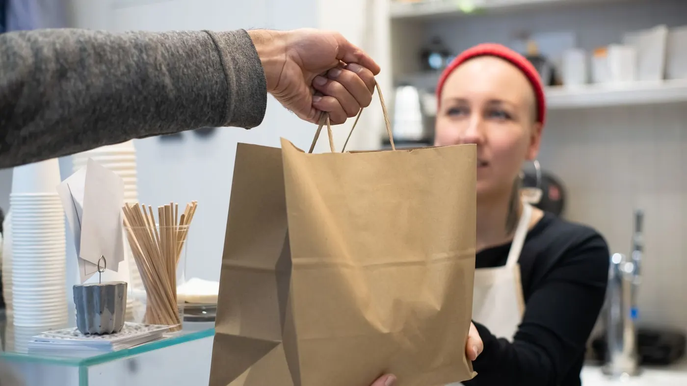 Restaurant worker in an apron handing a brown paper takeout bag across the counter to a customer